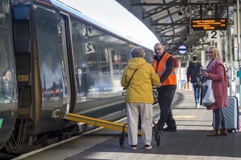 Staff assisting passenger with mobility aid onto train with ramp deployed at a station